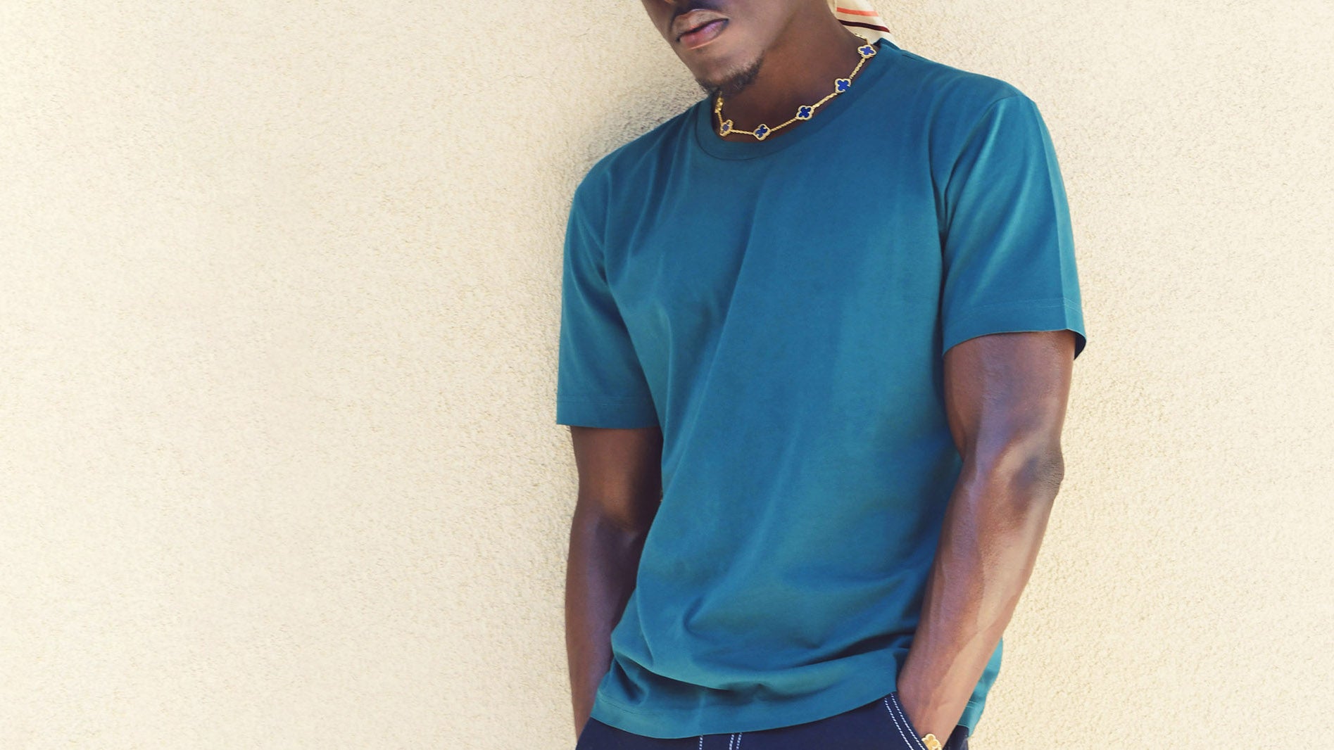 An African American Man leans against a beige wall. He wears the essential tee shirt in deep opal, a color similar to teal. It is a crewneck t-shirt with short sleeves.