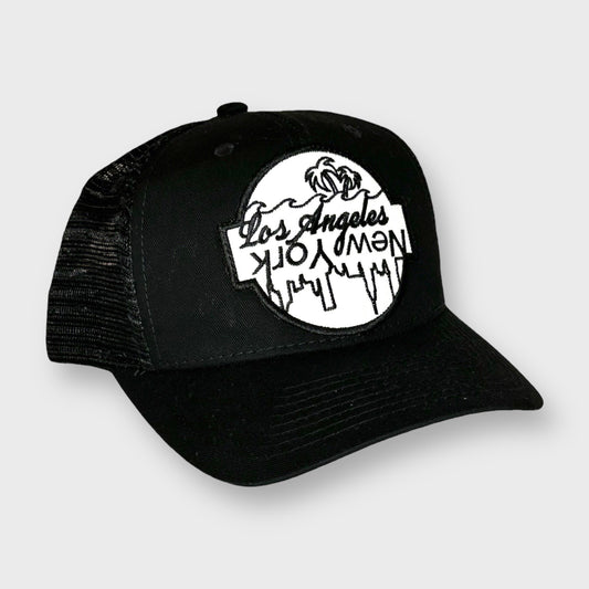 This is a black snapback hat with a white and black patch sewn onto the front. The design is a line drawing of palm trees and city scape with the words Los Angeles, and New York written in a reflection of one another. 