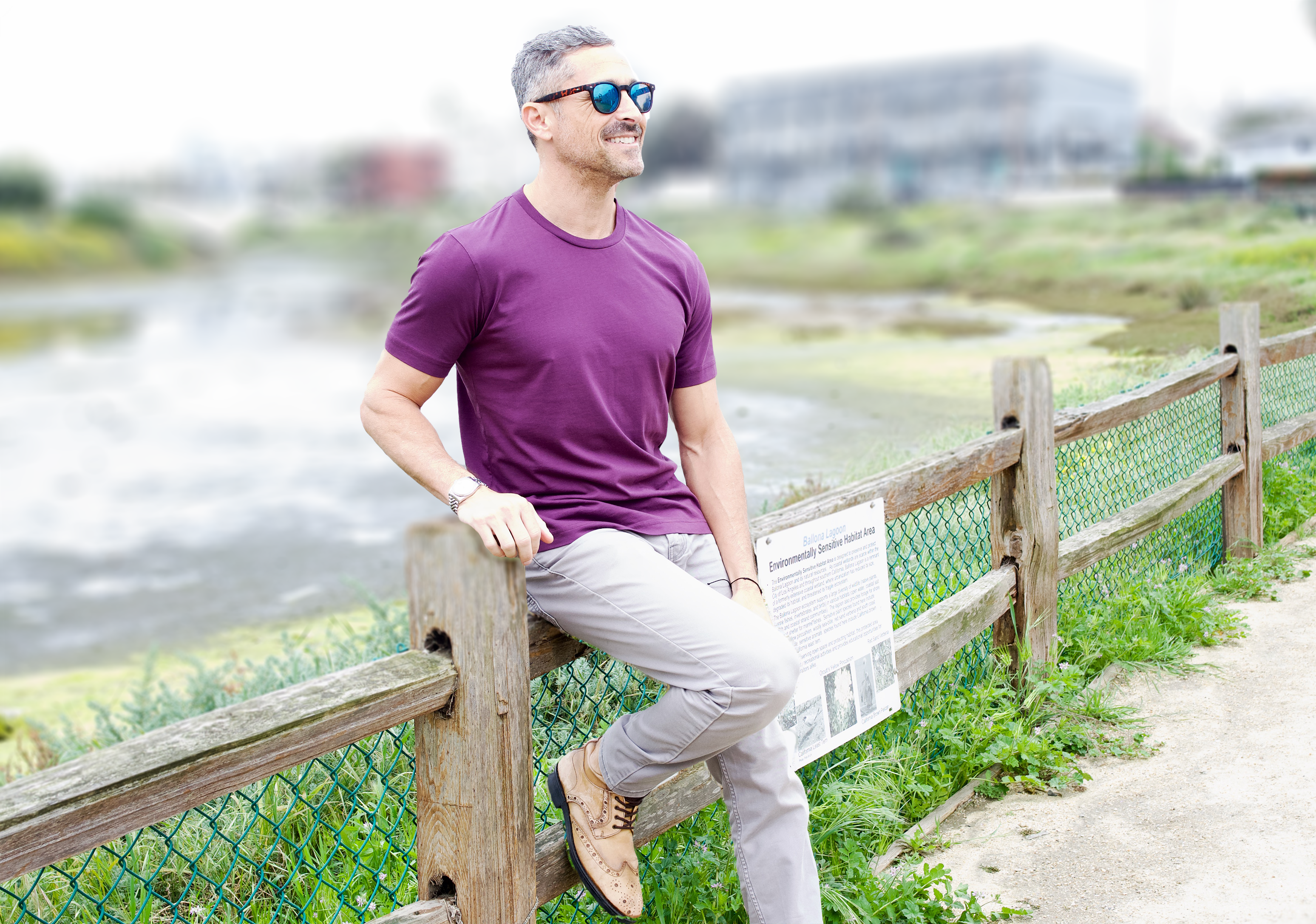 Model leans against a fence in Venice Canals in California. He wears sunglasses, a light aubergine crew neck tee shirt and grey pants. 